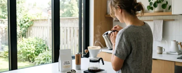 Une personne vue de dos verse de l'eau chaude dans un filtre à café, des grains de café frais disposés sur le comptoir d'une cuisine lumineuse contemporaine